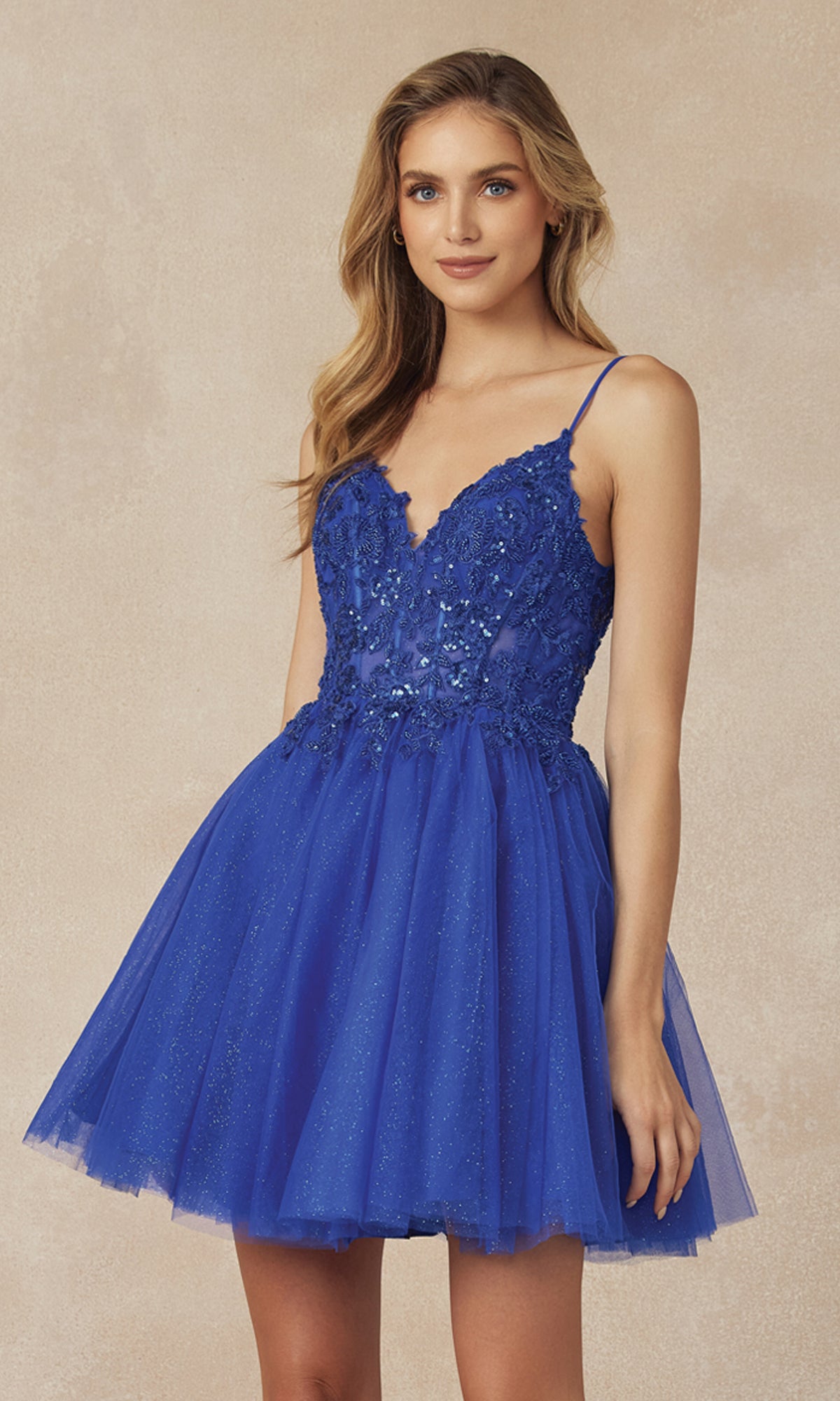 A woman wears the Sheer-Bodice Short A-Line Homecoming Dress 863, featuring a royal blue sleeveless V-neck corset-style bodice, tulle mini skirt, floral appliqué, and thin straps, standing against a beige background.